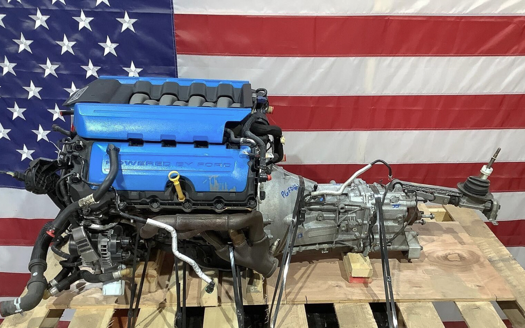 A Ford Coyote engine and transmission on a shipping pallet in front of an American flag