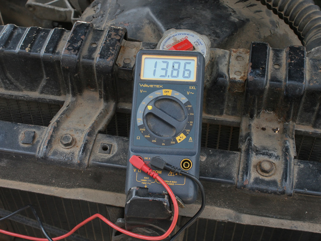 A digital multimeter in an engine bay reading 13.86 volts