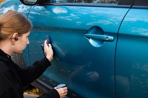 A woman using a waterless car wash spray and microfiber towel to detail an aqua car.