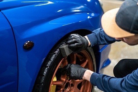 A technician wearing a two-tone flat brim hat applying tire shine to a blue car with bronze wheels.